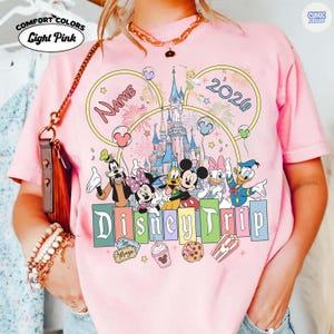 May include: Light pink Comfort Colors t-shirt with a Disney Trip design. The design features Disney characters, a castle, fireworks, and the year 2026. The word "Name" is also included in the design.