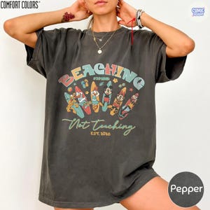 May include: Pepper-colored t-shirt with a colorful graphic that reads "BEACHING Not Teaching" above cartoon characters on surfboards. The shirt is from Comfort Colors and has "EST. 2026" printed below the text.