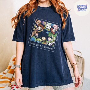 May include: A navy blue short-sleeve t-shirt featuring a square graphic print of various animated characters looking downward. Below the characters, the text reads "YOU'VE GOT A FRIEND IN ME" in white capital letters. The shirt has a crew neckline.