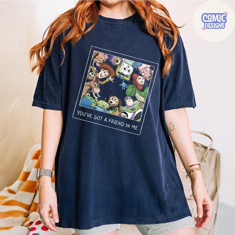 May include: A navy blue short-sleeve t-shirt featuring a square graphic print of various animated characters looking downward. Below the characters, the text reads "YOU'VE GOT A FRIEND IN ME" in white capital letters. The shirt has a crew neckline.