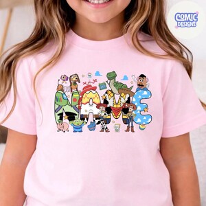 May include: A light pink short-sleeve t-shirt featuring a graphic print of the word NAME, where each letter is decorated with patterns and characters from the Toy Story franchise, including Woody, Buzz Lightyear, Jessie, and Rex.