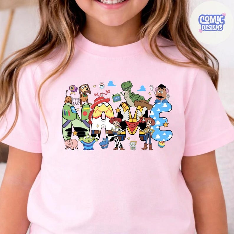 May include: A light pink short-sleeve t-shirt featuring a graphic print of the word NAME, where each letter is decorated with patterns and characters from the Toy Story franchise, including Woody, Buzz Lightyear, Jessie, and Rex.