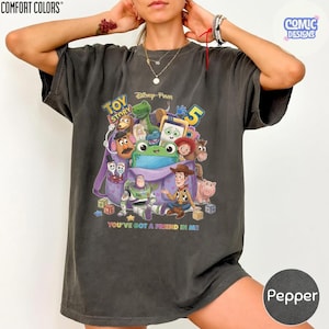 May include: A charcoal grey short-sleeve t-shirt featuring a colourful graphic print of Toy Story characters emerging from a purple backpack. The design includes the text Toy Story 5 and You've Got A Friend In Me, showcasing various animated figures.