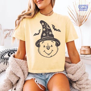 May include: A light yellow t-shirt featuring a cartoon bear wearing a witch hat with stars and bats. The shirt has a crew neck and short sleeves. The design is in black and white.
