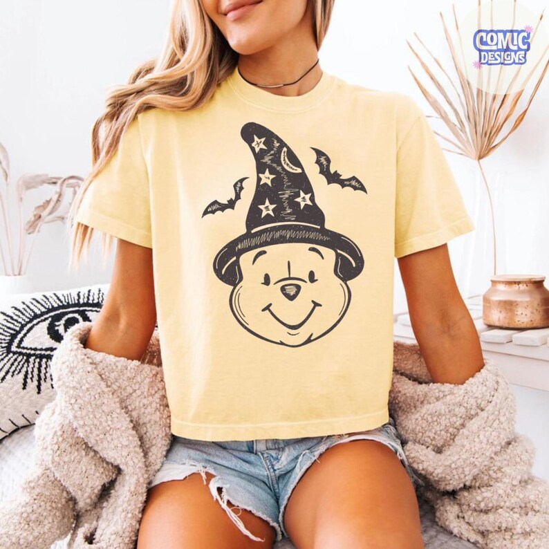 May include: A light yellow t-shirt featuring a cartoon bear wearing a witch hat with stars and bats. The shirt has a crew neck and short sleeves. The design is in black and white.