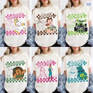 May include: A collection of six off-white t-shirts featuring various cartoon characters from Toy Story, including Buttercup, Anny, RC Car, Mr. Pricklepants, Barbie, and Bunny. Each shirt displays a character graphic with a checkered border.