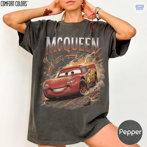 Pode incluir: T-shirt cinza escuro com um desenho de Rel&acirc;mpago McQueen do filme Carros. O design inclui o nome "MCQUEEN" e o n&uacute;mero "95" no carro de corrida vermelho. A camisa &eacute; da marca Comfort Colors.