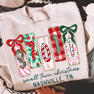 May include: A cream-colored sweatshirt with a festive Christmas design. The design features the numbers "2011" in a colorful, decorative style with bows and patterns. The text "small town christmas NASHVILLE, TN" is printed below the numbers.