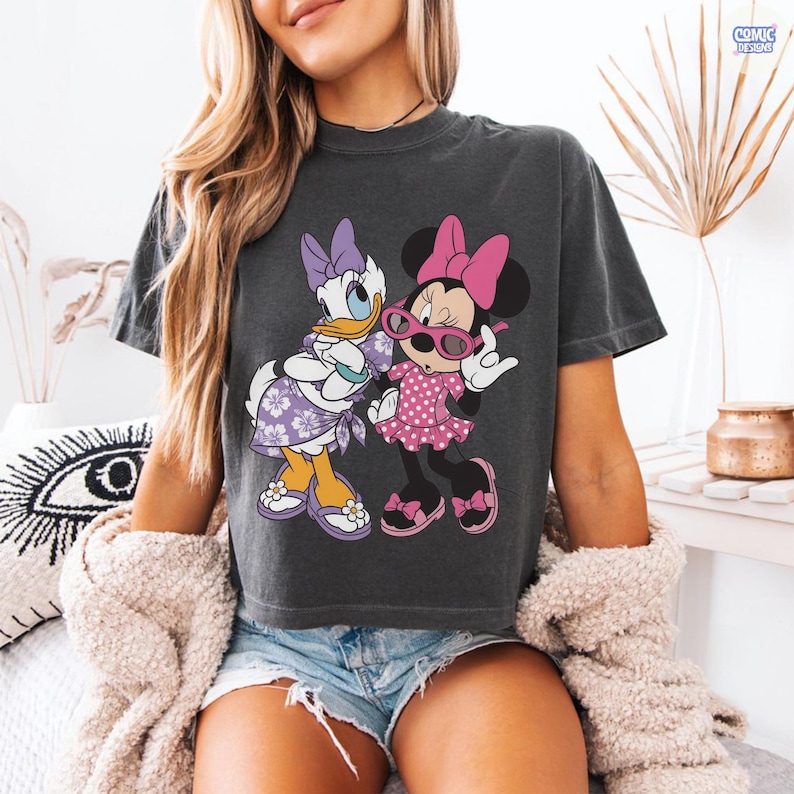 Puede incluir: Una camiseta de manga corta gris carb&oacute;n con un estampado gr&aacute;fico de Daisy Duck y Minnie Mouse. Daisy lleva un vestido y un lazo con flores moradas, mientras que Minnie lleva un vestido de lunares rosa, un lazo rosa y gafas rosas.