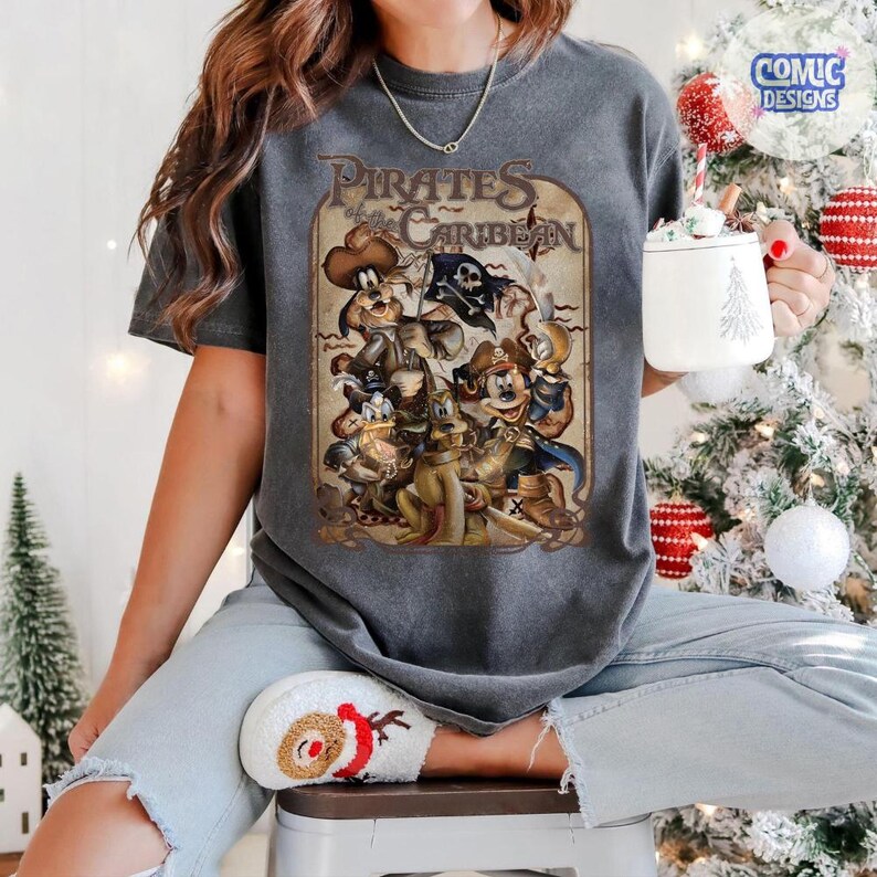 May include: Dark gray t-shirt featuring a vintage-style graphic of Disney characters as pirates, with the text "Pirates of the Caribbean". The design includes Mickey Mouse, Donald Duck, and Goofy in pirate attire. The shirt is worn by a person.