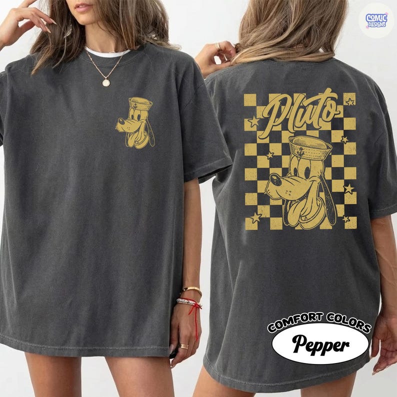May include: Dark grey t-shirt with a gold Pluto graphic on the front and back. The back design features Pluto in a sailor hat, the name "Pluto", and a gold and black chequered pattern. The shirt is labelled "Comfort Colors Pepper".