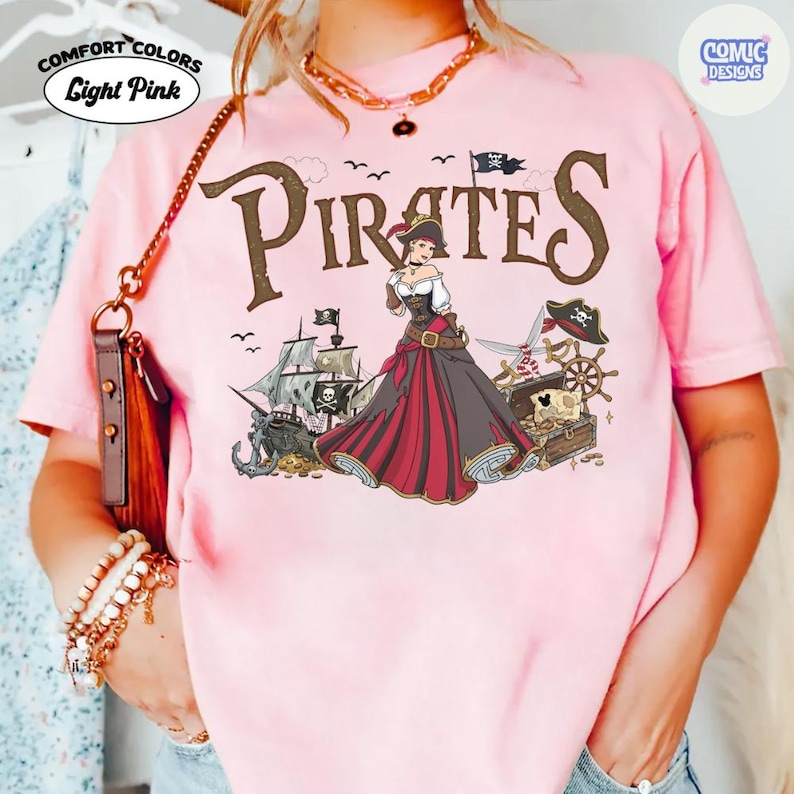 May include: Light pink t-shirt with a pirate-themed graphic. The design features the word "Pirates" in a distressed font, a pirate ship, and a female pirate character in a red and black dress. The shirt also has the words "Comfort Colors" and "Light Pink".