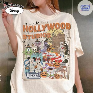 K&ouml;nnte beinhalten: Ein elfenbeinfarbenes T-Shirt mit einem Grafikdruck, der den Text "HOLLYWOOD STUDIOS" und "ROCKERS" enth&auml;lt. Das Design zeigt verschiedene Zeichentrickfiguren in Halloween-Kost&uuml;men, K&uuml;rbisse, Spinnennetze und die Silhouette eines Geb&auml;udes.