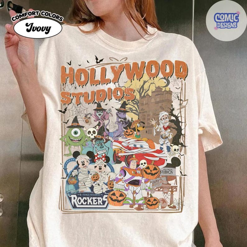 K&ouml;nnte beinhalten: Ein elfenbeinfarbenes T-Shirt mit einem Grafikdruck, der den Text "HOLLYWOOD STUDIOS" und "ROCKERS" enth&auml;lt. Das Design zeigt verschiedene Zeichentrickfiguren in Halloween-Kost&uuml;men, K&uuml;rbisse, Spinnennetze und die Silhouette eines Geb&auml;udes.