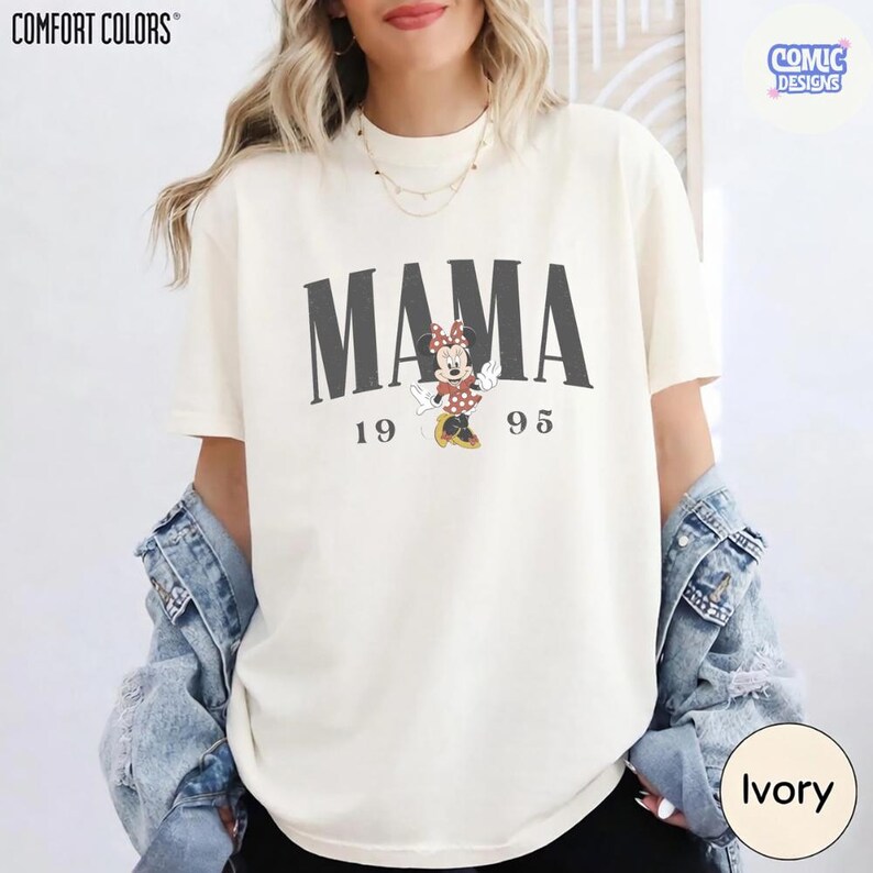 K&ouml;nnte beinhalten: Elfenbeinfarbenes Comfort Colors T-Shirt mit dem Wort "MAMA" in dunklen, grauen Buchstaben. Eine Minnie Mouse-Grafik befindet sich darunter, mit den Zahlen "19" und "95" auf beiden Seiten. Das Shirt geh&ouml;rt zur Comic Designs Kollektion.