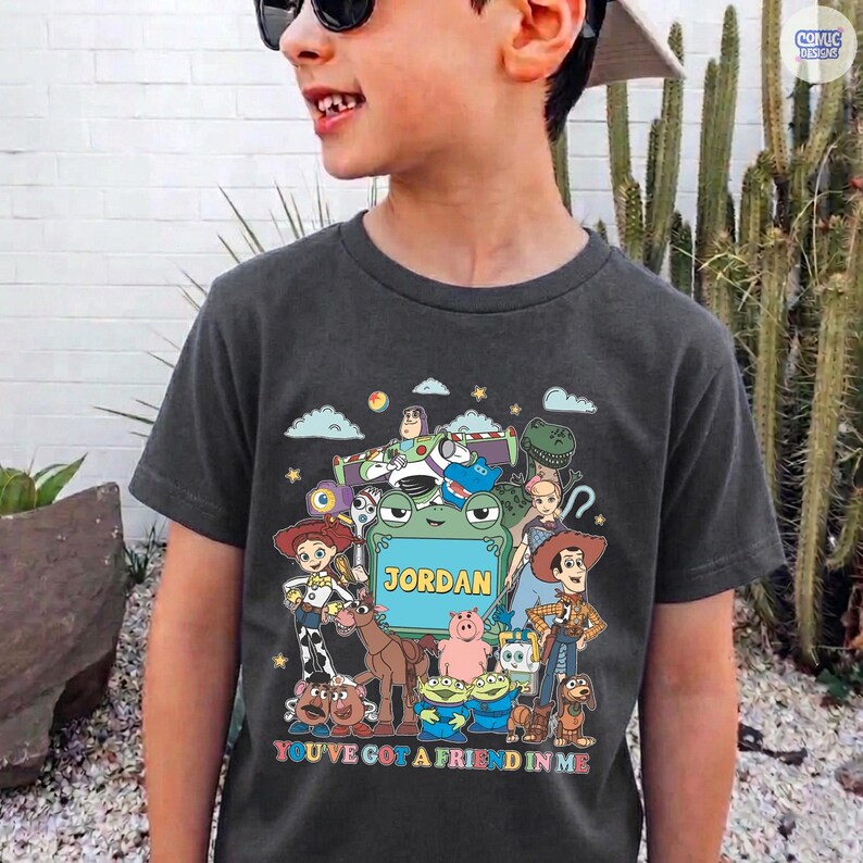 May include: A dark gray short-sleeve t-shirt featuring a colorful graphic print of various animated characters from Toy Story surrounding a central green frog frame with the name JORDAN printed in yellow text and the phrase YOU'VE GOT A FRIEND IN ME.