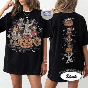 Op de afbeelding: Een zwart t-shirt met een grote afbeelding op de voorkant van stripfiguren in Halloween-kostuums rond een pompoen, en een verticale afbeelding op de achterkant met dezelfde figuren, inclusief de tekst Comic Designs.