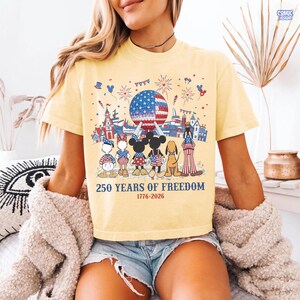 May include: A pale yellow t-shirt featuring a graphic of cartoon characters standing before a large sphere with an American flag pattern, fireworks, and the text 250 YEARS OF FREEDOM 1776-2026. The design includes various patriotic motifs and icons.