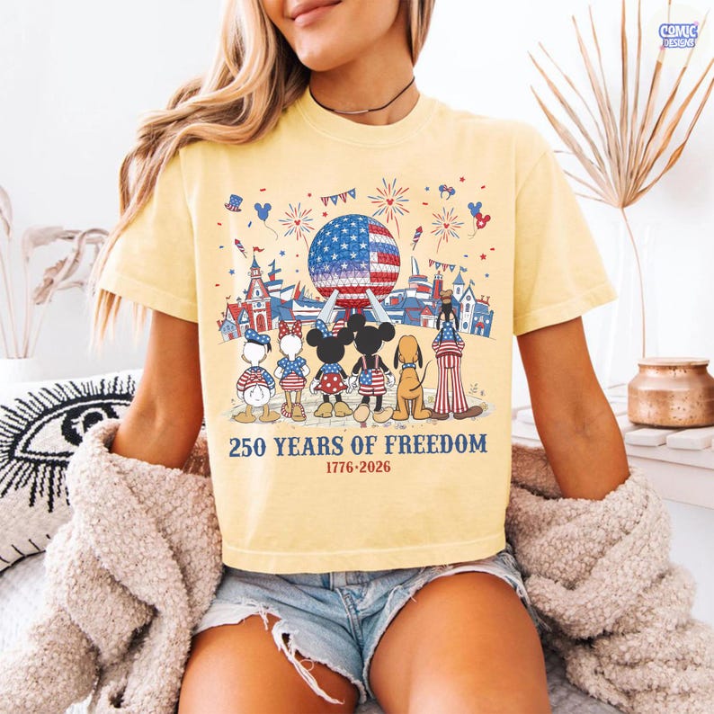 May include: A pale yellow t-shirt featuring a graphic of cartoon characters standing before a large sphere with an American flag pattern, fireworks, and the text 250 YEARS OF FREEDOM 1776-2026. The design includes various patriotic motifs and icons.