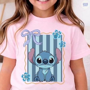 May include: Pink t-shirt featuring a cartoon image of Stitch from Lilo & Stitch. The design includes a blue Stitch character, a blue bow, and blue floral accents on a striped background. The shirt is made of a soft material.