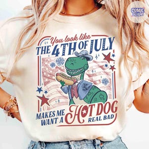 May include: Cream-colored t-shirt featuring a green dinosaur wearing a hat and suit, holding a hot dog. The shirt has text that reads "You look like the 4th of July makes me want a hot dog real bad" with patriotic stars and stripes.