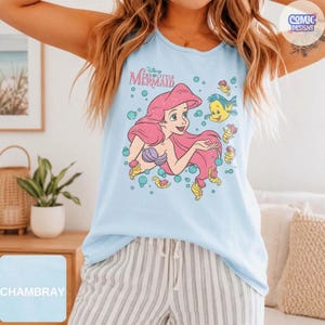 May include: A light blue sleeveless tank top featuring a graphic print of Ariel from The Little Mermaid with her long pink hair, surrounded by bubbles and the yellow fish Flounder. The text Disney The Little Mermaid is printed above the character.