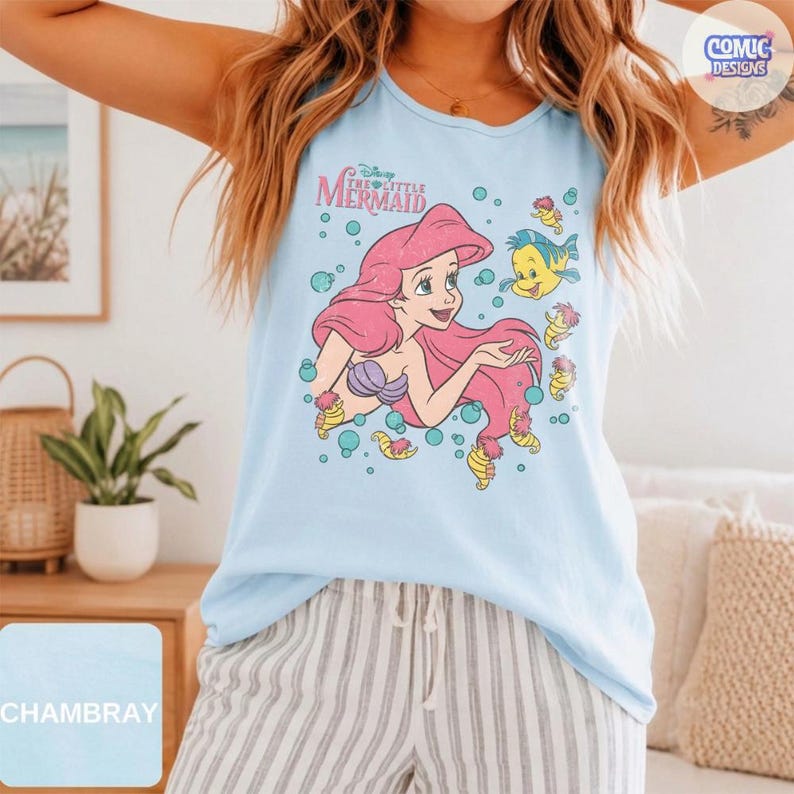 May include: A light blue sleeveless tank top featuring a graphic print of Ariel from The Little Mermaid with her long pink hair, surrounded by bubbles and the yellow fish Flounder. The text Disney The Little Mermaid is printed above the character.