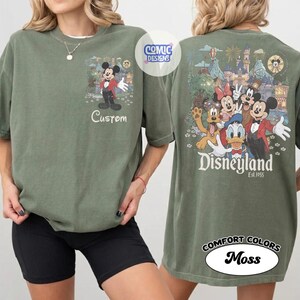 May include: An olive green t-shirt featuring a small graphic of Mickey Mouse on the front chest with the text Custom. The back displays a large, colorful illustration of Mickey, Minnie, Donald, Goofy, and Pluto with the text Disneyland Est. 1955.