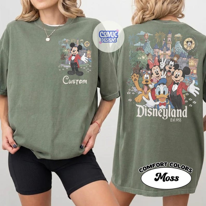 May include: An olive green t-shirt featuring a small graphic of Mickey Mouse on the front chest with the text Custom. The back displays a large, colorful illustration of Mickey, Minnie, Donald, Goofy, and Pluto with the text Disneyland Est. 1955.