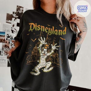 Może przedstawiać: Ciemnoszary t-shirt z grafiką przedstawiającą postać Goofy'ego owiniętego w bandaże mumii na tle zarysu zamku. Słowo Disneyland jest nadrukowane zieloną, ściekającą czcionką nad postacią, w otoczeniu małych pomarańczowych sylwetek nietoperzy.