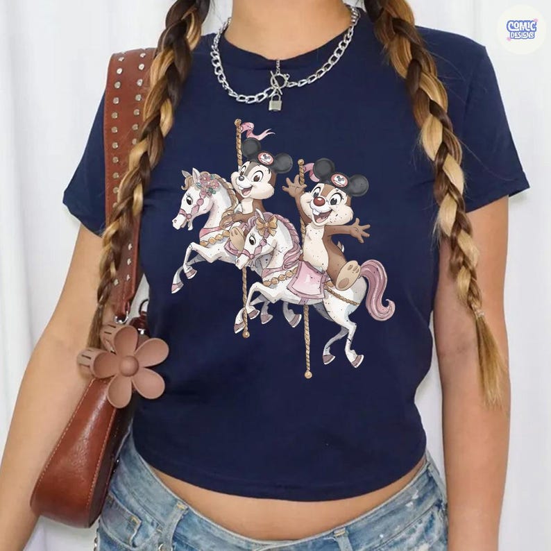 May include: Navy blue t-shirt featuring a carousel design with cartoon characters riding white horses. The graphic includes pink and gold accents. The shirt has short sleeves and a crew neck. The Comic Designs logo is visible in the upper right corner.