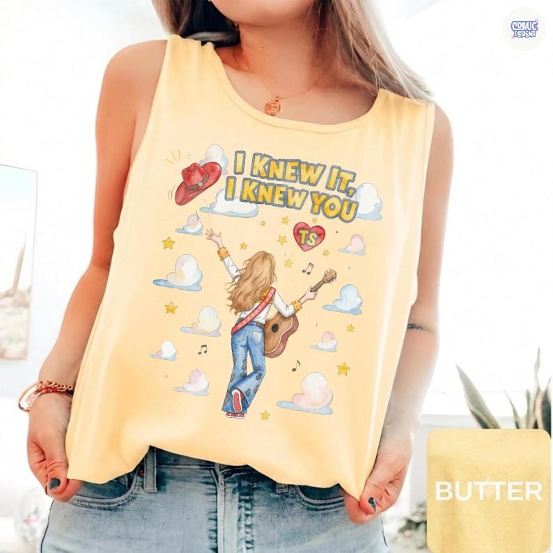 May include: A pale yellow sleeveless tank top featuring a graphic illustration of a person with long hair playing an acoustic guitar. The design includes clouds, stars, a red cowboy hat, a heart with the letters TS, and the text I KNEW IT, I KNEW YOU.
