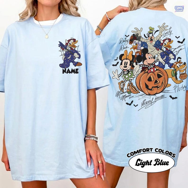 May include: A light blue t-shirt featuring a small graphic of Donald Duck in a star-patterned costume on the front chest above the word NAME. The back displays a large Halloween-themed illustration of Mickey, Minnie, Goofy, Pluto, and Daisy Duck.