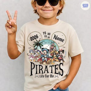 May include: Beige t-shirt featuring a pirate-themed design with Mickey Mouse and friends. The design includes the word "PIRATES" in a bold font, the text "Life for Me", and the year "2026". The shirt has a crew neck and short sleeves.