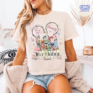 May include: A cream-colored t-shirt featuring a colorful graphic of Disney characters, the number 5, and the name "Amelia" in a Mickey Mouse ear design. The words "Birthday Squad" are printed below the characters.