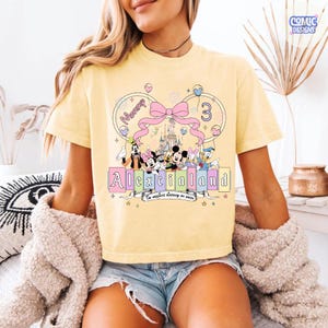 Op de afbeelding: Lichtgeel T-shirt met een kleurrijk grafisch ontwerp. Het ontwerp bevat de tekst "Alexcialand" en "The happiest birthday on earth" met Disney-figuren en een kasteel. Een roze strik en het nummer 3 zijn ook inbegrepen.