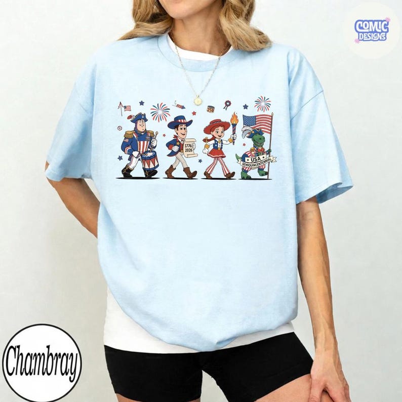 May include: Light blue t-shirt featuring a patriotic cartoon parade with characters holding a drum, a scroll, a torch, and an American flag. The shirt has the text "Comic Designs" and "Chambray".
