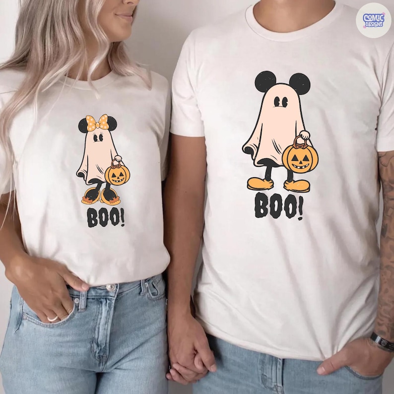 May include: White t-shirts with a Halloween-themed design. The design features a ghost with Mickey Mouse ears holding a jack-o'-lantern and the word "BOO!". One shirt has a bow on the ghost's head. The shirts are worn by two people.