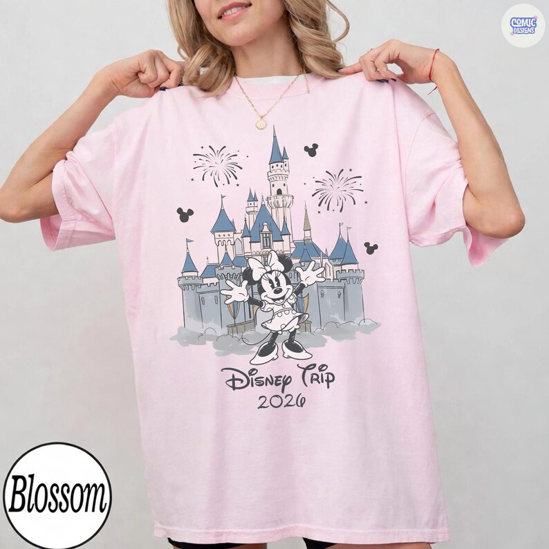 May include: Pink t-shirt featuring a graphic of Minnie Mouse in front of a castle with fireworks and the text "Disney Trip 2026". The shirt has short sleeves and a round neckline.