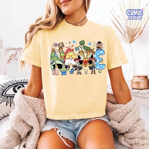 May include: A pale yellow short-sleeve t-shirt featuring a large graphic print of the word NAME. The letters are filled with patterns and illustrations of various animated toy characters, including a cowboy, a space ranger, and a dinosaur.