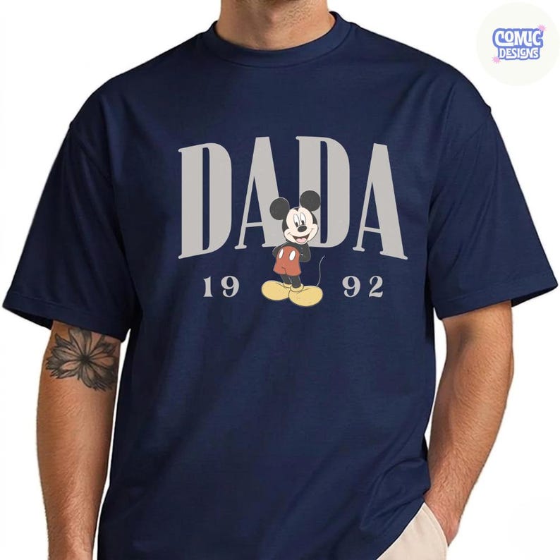 K&ouml;nnte beinhalten: Marineblaues T-Shirt mit dem Wort "DADA" in grauen Blockbuchstaben und einer Mickey Mouse-Grafik. Die Zahlen "19" und "92" stehen darunter. Das T-Shirt hat kurze &Auml;rmel und einen Rundhalsausschnitt.