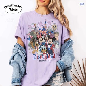 Puede incluir: Una camiseta de color violeta claro con un gr&aacute;fico de Mickey Mouse, Minnie Mouse, Donald Duck y Pluto vistiendo ropa patri&oacute;tica roja, blanca y azul. El dise&ntilde;o incluye un castillo, fuegos artificiales y el texto Disneyland America 250.