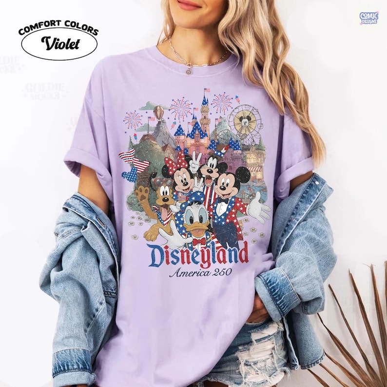 Puede incluir: Una camiseta de color violeta claro con un gr&aacute;fico de Mickey Mouse, Minnie Mouse, Donald Duck y Pluto vistiendo ropa patri&oacute;tica roja, blanca y azul. El dise&ntilde;o incluye un castillo, fuegos artificiales y el texto Disneyland America 250.