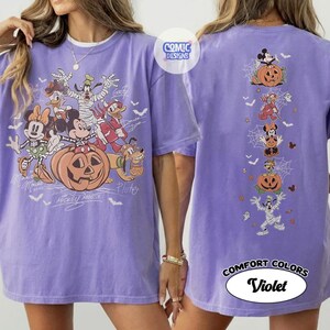 Op de afbeelding: Een violet t-shirt met een grote afbeelding op de voorkant van Disney-figuren in Halloween-kostuums met een pompoen en een verticale afbeelding op de achterkant, met de tekst COMIC DESIGNS en COMFORT COLORS Violet.