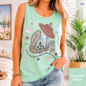 Puede incluir: Una camiseta sin mangas de color verde menta claro con un estampado gr&aacute;fico de una bota vaquera con patr&oacute;n de manchas de vaca, un sombrero marr&oacute;n y bloques apilados con el texto "13". El dise&ntilde;o incluye flores, estrellas y cuentas.