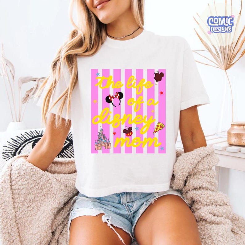 Puede incluir: Camiseta blanca con dise&ntilde;o de rayas rosas y blancas. La camiseta presenta el texto "The life of a Disney mom" en amarillo, junto con gr&aacute;ficos tem&aacute;ticos de Disney como orejas de Mickey Mouse, un castillo y pizza.