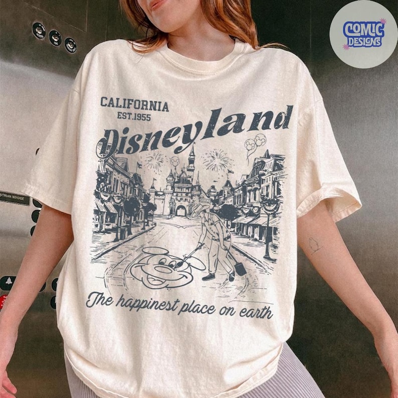 May include: Cream-coloured t-shirt with a Disneyland graphic. The design includes the text "CALIFORNIA EST.1955 Disneyland" and a detailed illustration of a street scene with fireworks and Mickey Mouse. The phrase "The happiest place on earth" is also printed on the shirt.