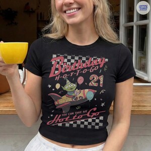 May include: Black short-sleeved t-shirt with a graphic featuring an alien character in a rocket ship, the text "Birthday H-O-T-T-O-GO 21" and "You can take me Hot to Go". The design includes checkered patterns and stars.
