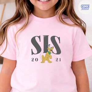 K&ouml;nnte beinhalten: Hellrosa T-Shirt mit dem Wort "SIS" in Dunkelgrau, einer Cartoon-Pluto-Grafik und den Zahlen "20" und "21". Das T-Shirt hat einen Rundhalsausschnitt und kurze &Auml;rmel. Ein lustiges, l&auml;ssiges Oberteil.