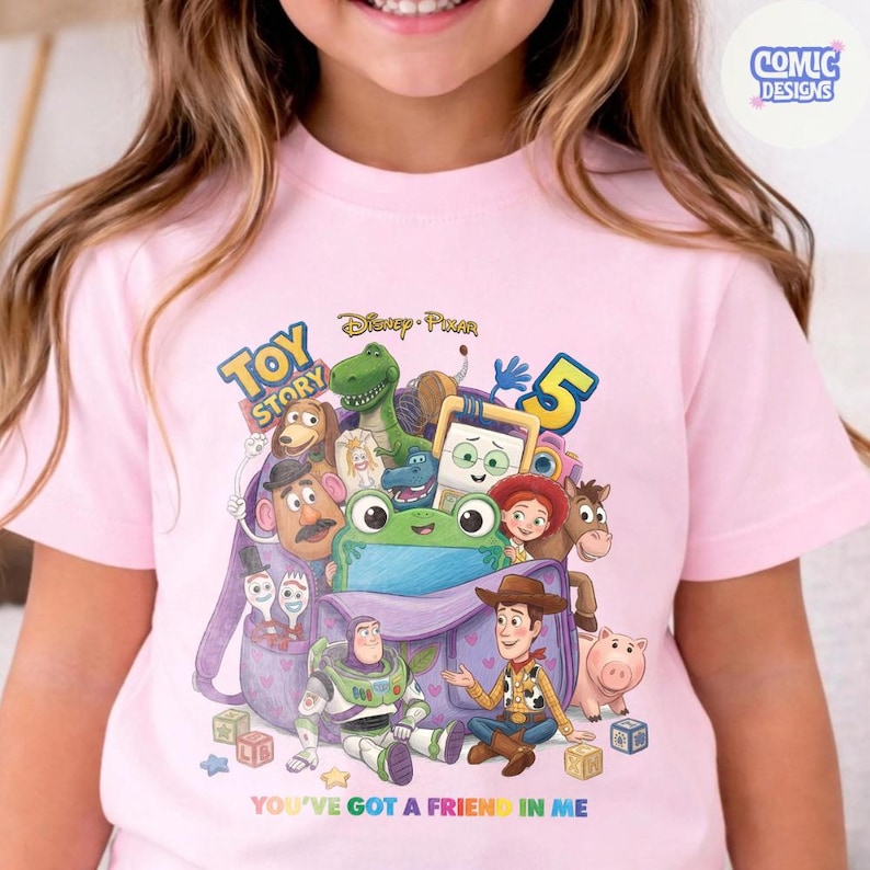 May include: A light pink t-shirt featuring a graphic print of various Toy Story characters emerging from a purple backpack. The design includes the text Toy Story, the number 5, and the phrase You've Got A Friend In Me printed at the bottom.